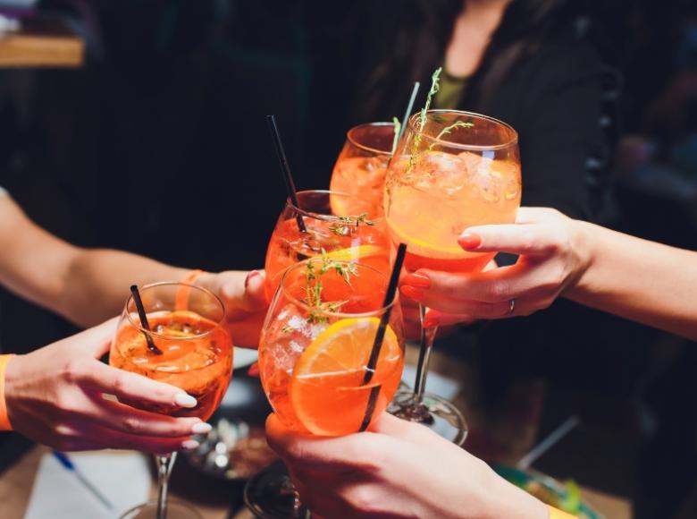 Persone brindano con cocktail arancioni, decorati con fette d'arancia e rametti di erbe.